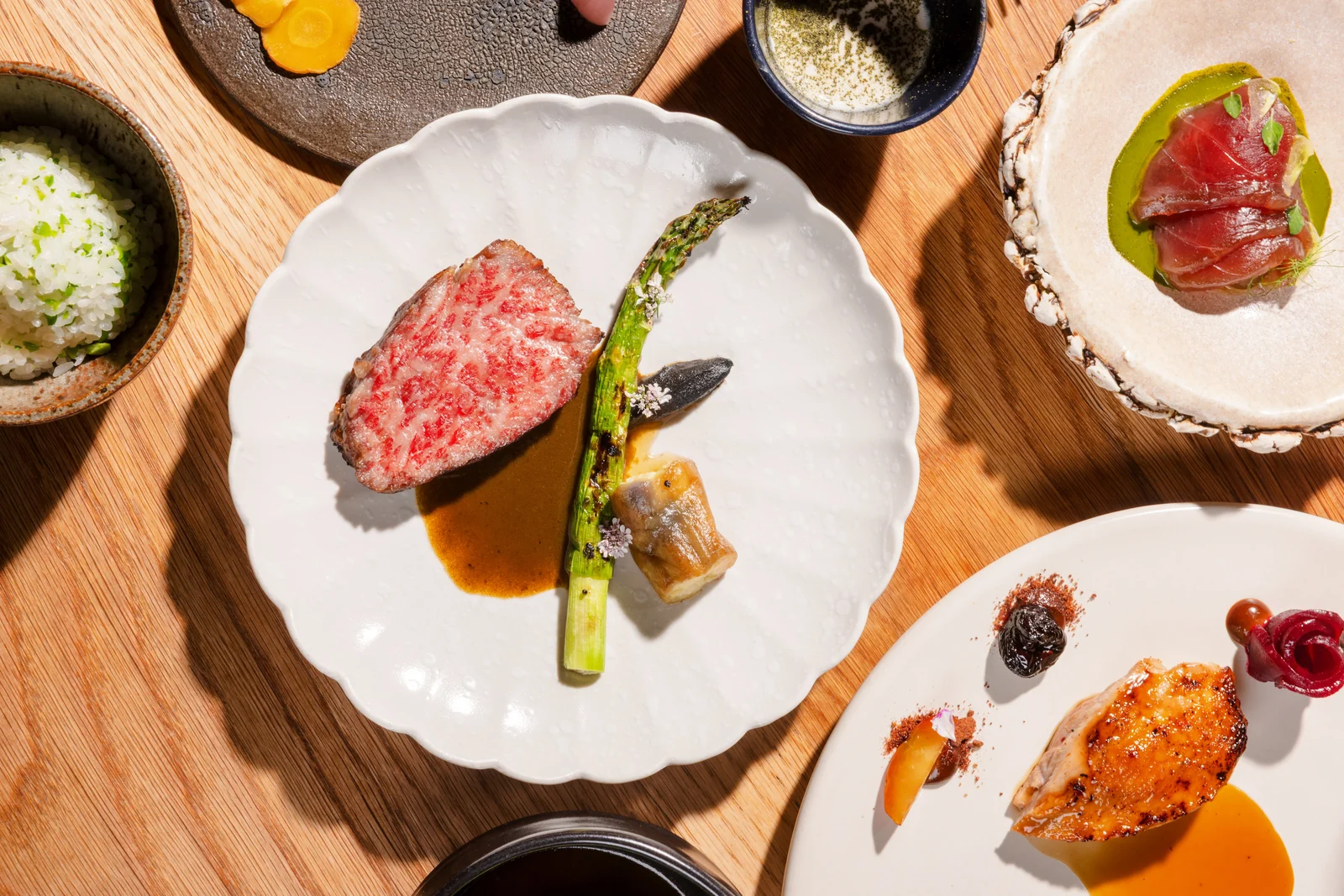 Plated dishes on a wooden pass in daylight: sliced wagyu with grilled asparagus, tuna crudo in herb oil, herbed rice, and a glazed poultry course.
